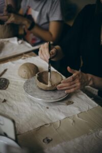 A close-up of hands shaping a clay bowl with a paintbrush in a pottery class setting.